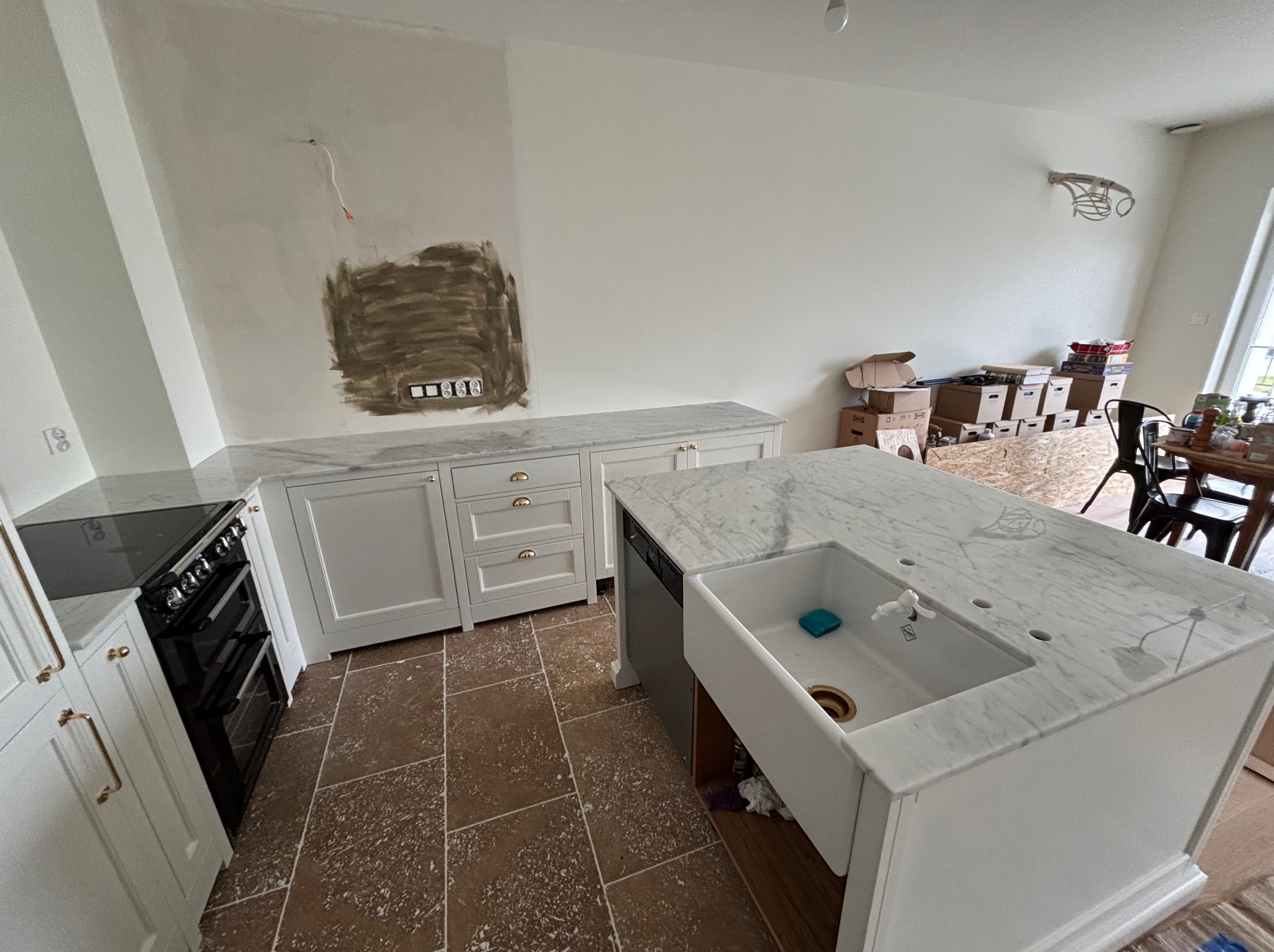 Kuchnia w trakcie remontu: białe szafki z marmurowym blatem, brązowa podłoga, ściana z widocznymi śladami po instalacjach. Widoczny fragment wyspy kuchennej z umywalką.