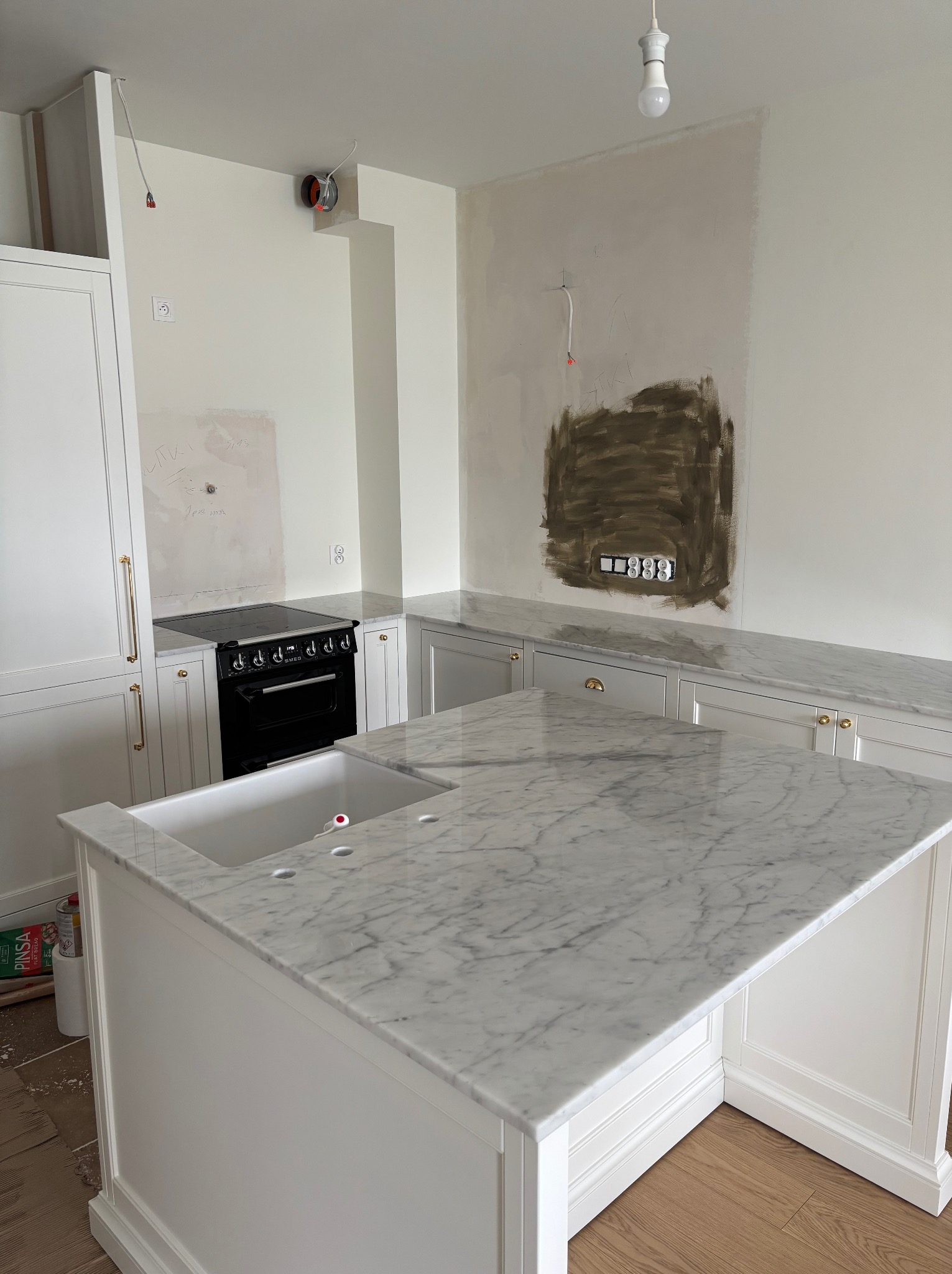 Biała kuchnia w trakcie wykończenia: marmurowy blat z wbudowanym zlewem, czarna kuchenka, niedokończone ściany. Widoczne kable i puszki elektryczne.