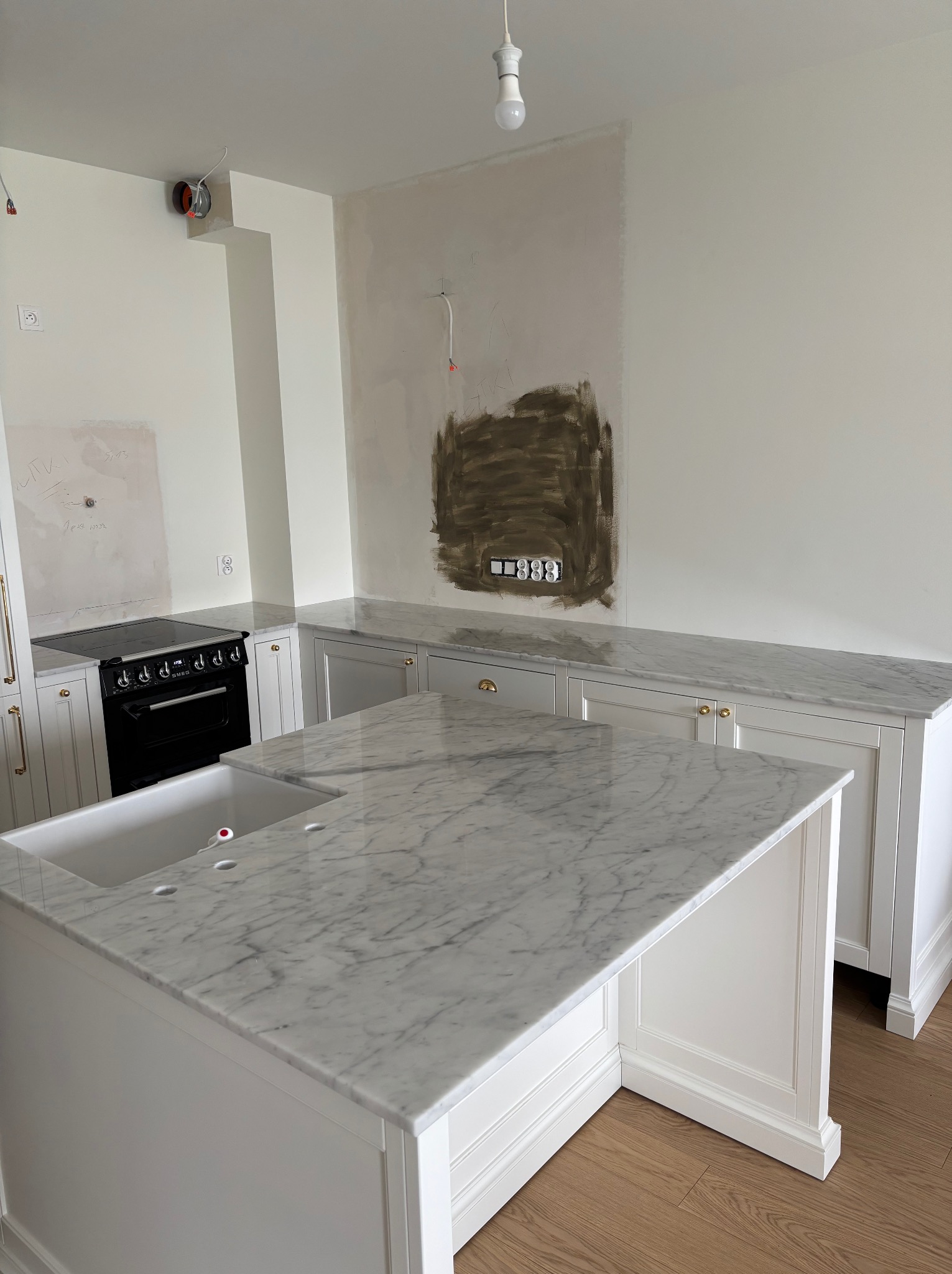 Biała kuchnia w trakcie montażu: marmurowy blat, wyspa z umywalką, ściany częściowo wykończone, widoczne instalacje elektryczne.