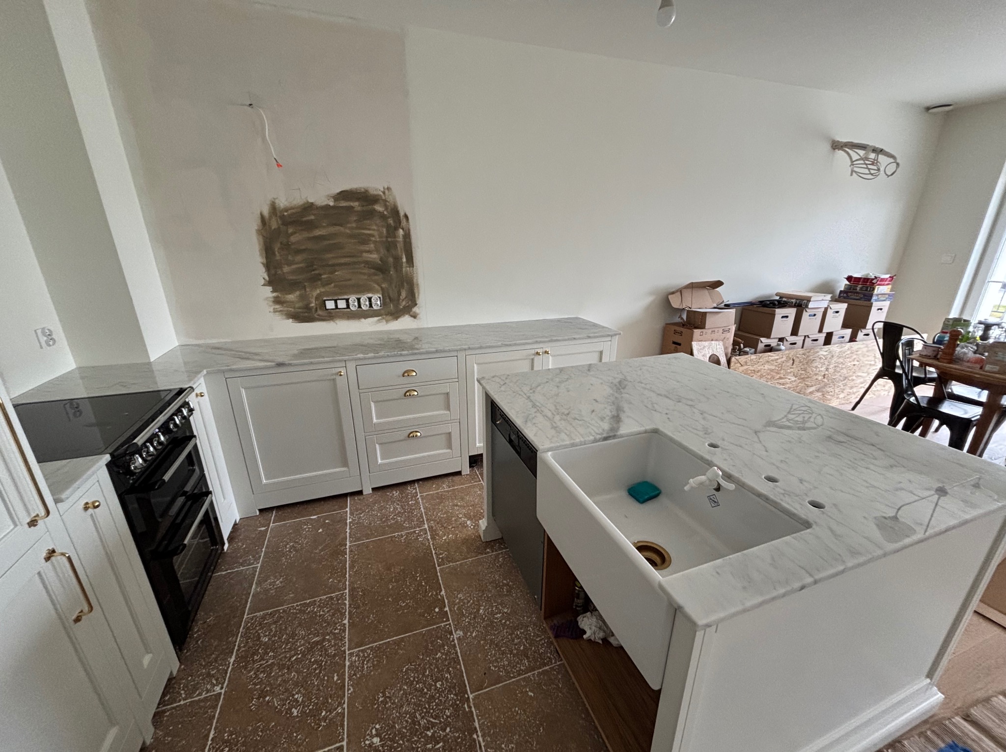 Jasna kuchnia w trakcie wykończenia: białe szafki z marmurowym blatem, wyspa z ceramicznym zlewem farmerskim, brązowa podłoga z płytek. Ściany w trakcie malowania.
