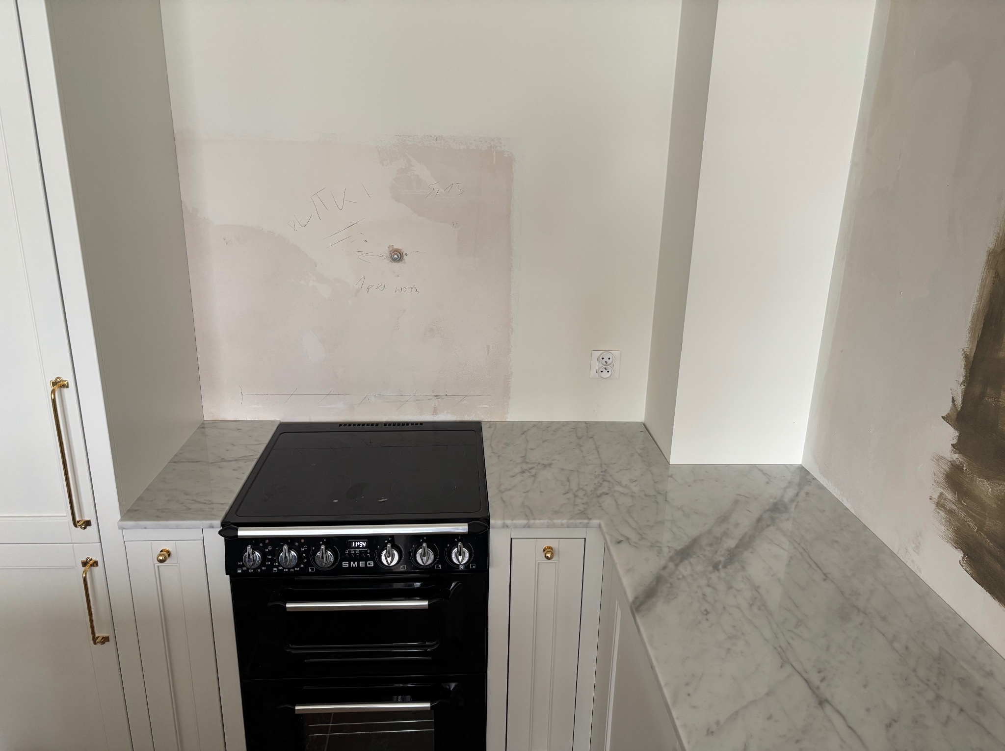 Marmurowy blat kuchenny obok czarnej kuchenki SMEG, fragment remontu kuchni z widocznymi śladami prac na ścianie i złotymi uchwytami szafek.