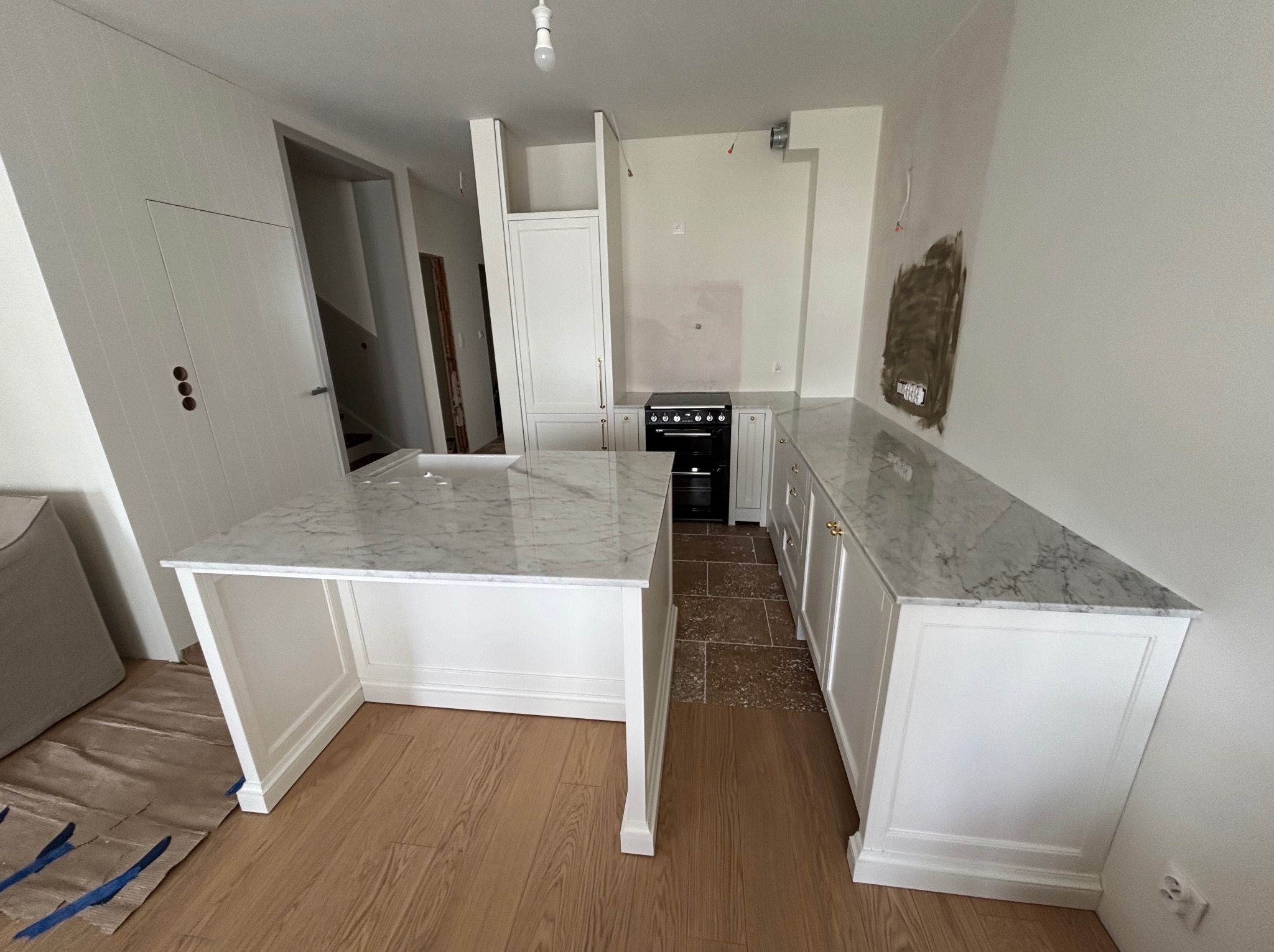 Jasna kuchnia w trakcie wykańczania z marmurowym blatem, widoczne zabezpieczenia podłogi. Na ścianie ślady po instalacji. W tle czarny piekarnik i białe szafki.