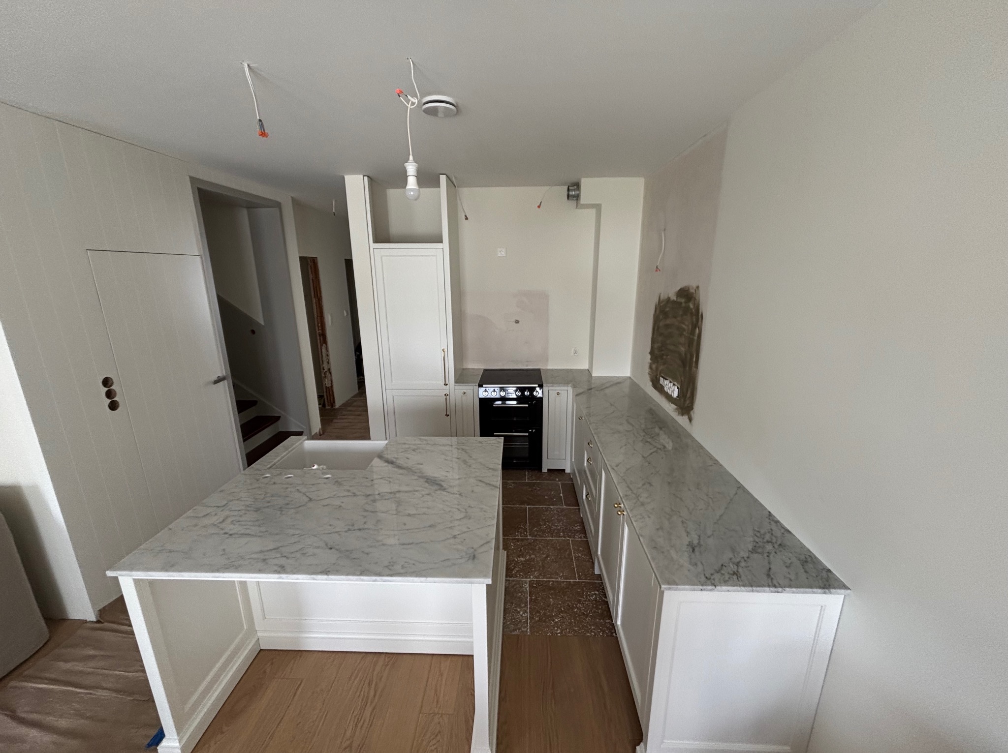 Jasna kuchnia w trakcie montażu: wyspa z marmurowym blatem, zabudowa z białymi frontami i kamienny blat wzdłuż ściany. Widoczne niedokończone prace wykończeniowe.