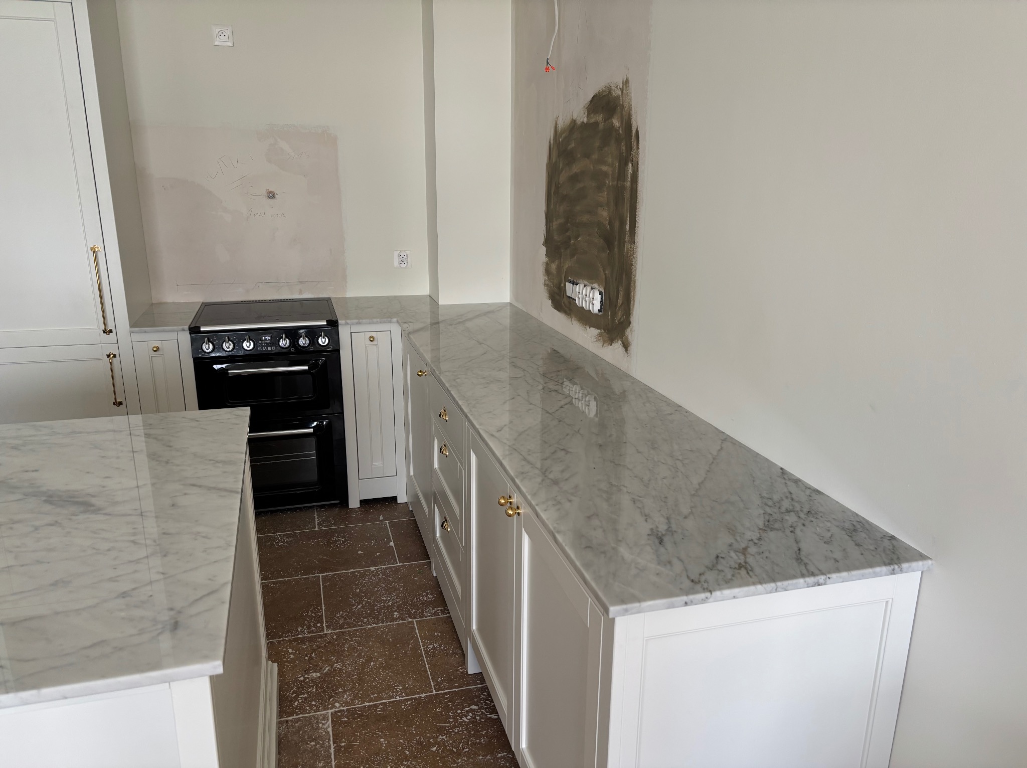 Biały blat kamienny z szarymi żyłkami na szafkach kuchennych z białymi frontami i złotymi uchwytami. W tle czarny piekarnik i fragment ściany z instalacją elektryczną.