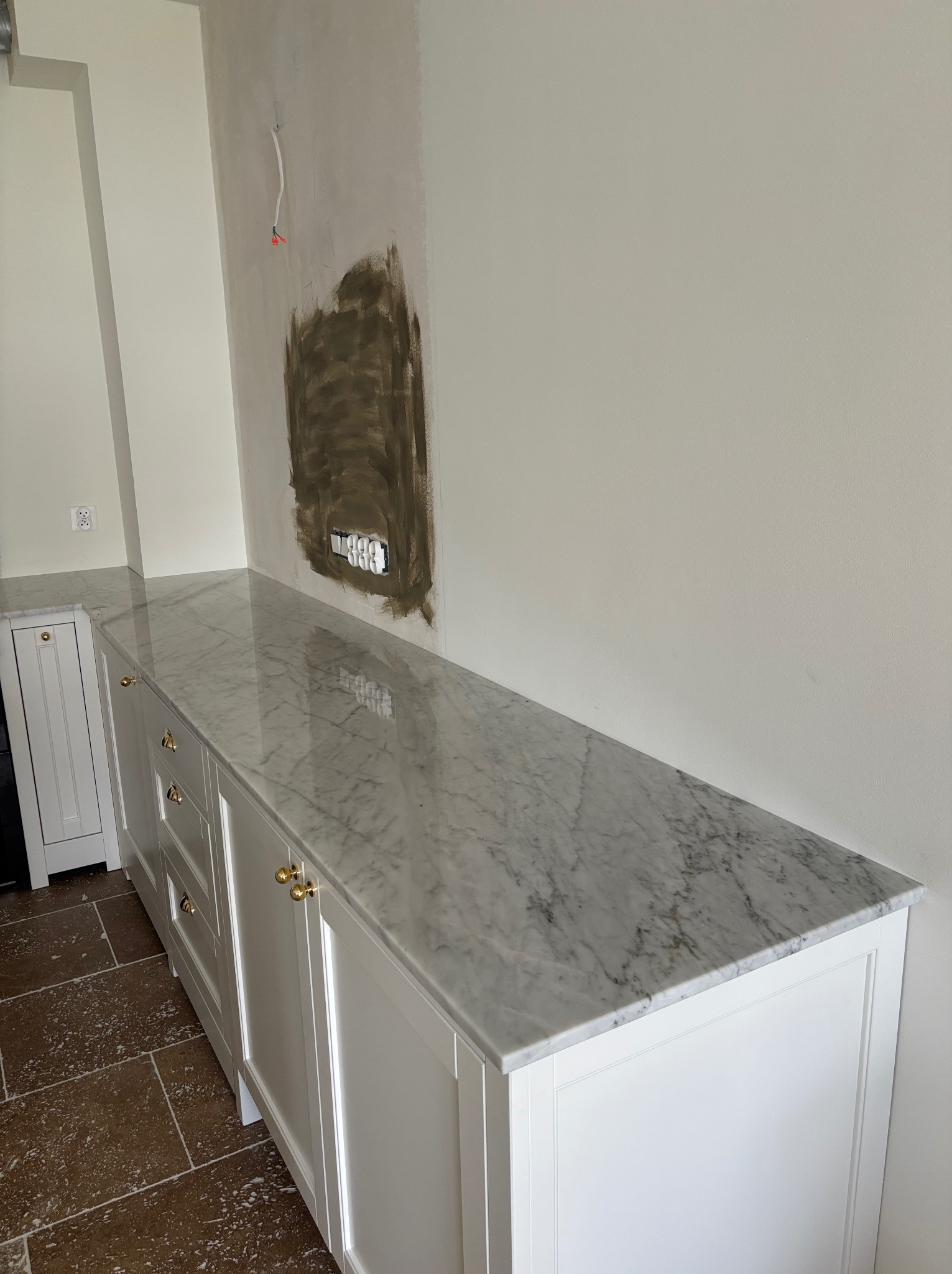 Biała szafka z marmurowym blatem w trakcie montażu. Widoczne niedokończone prace na ścianie i wystające kable. Złote uchwyty dodają elegancji.