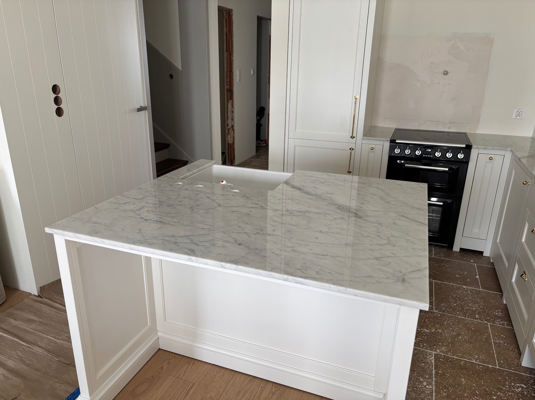 Kuchenna wyspa z blatem z białego marmuru, z widocznym zlewem. W tle białe szafki, czarny piekarnik Smeg i fragment brązowej podłogi. Wnętrze w trakcie wykończenia.