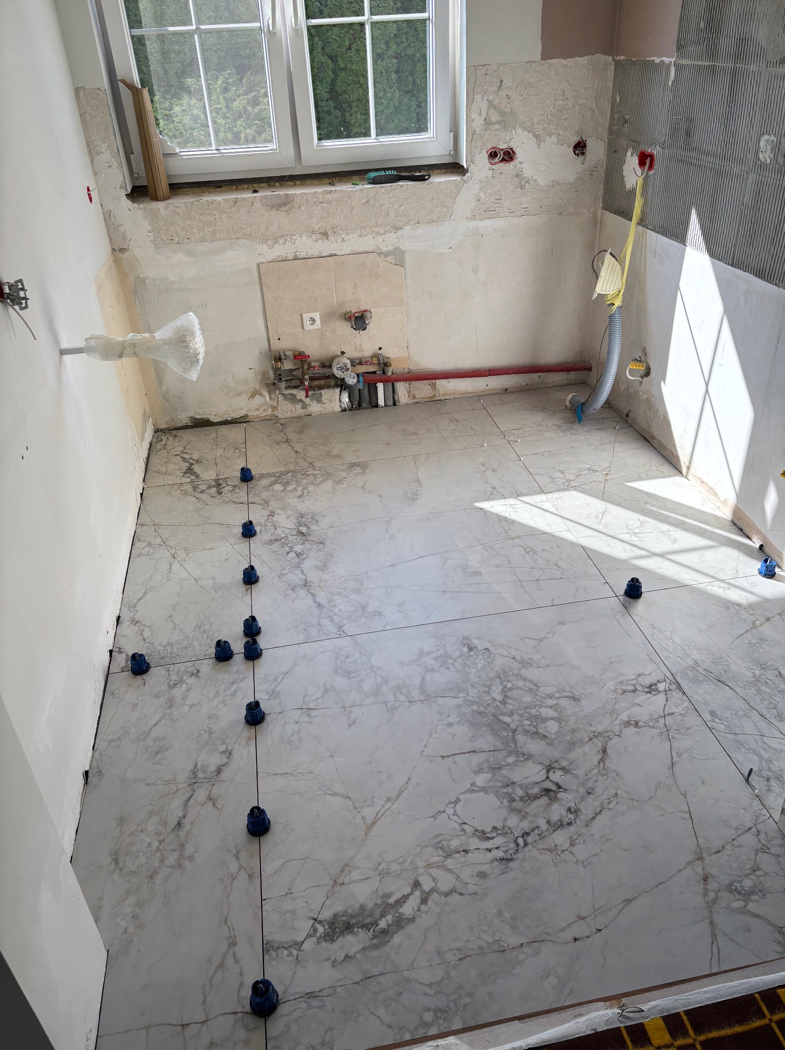 Układanie marmurowych płytek podłogowych w kuchni, widoczne niebieskie klipsy poziomujące. Częściowo wykończone ściany, okno w tle.