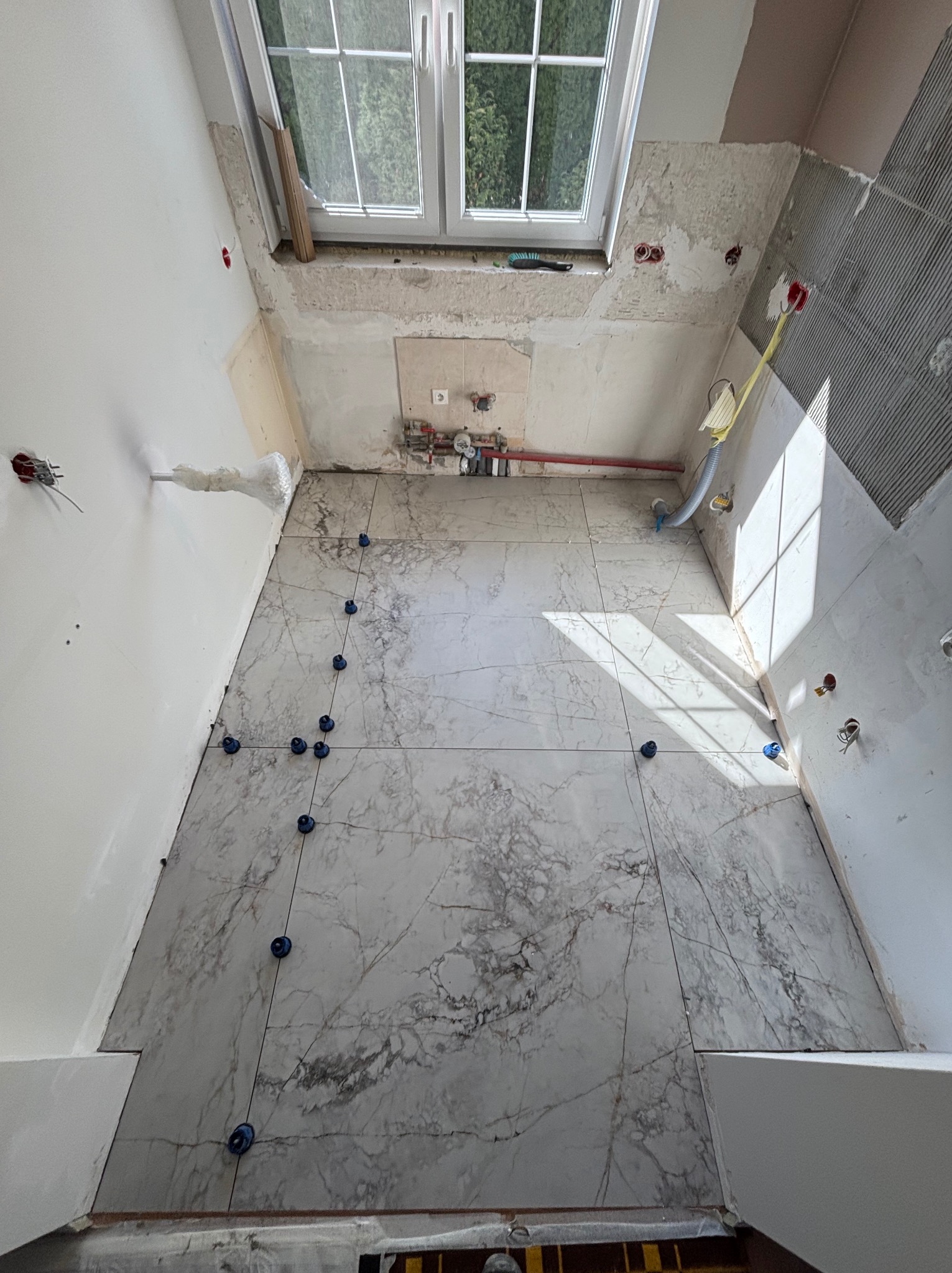 Montaż marmurowych płytek podłogowych w łazience z oknem dachowym, z użyciem systemu poziomowania płytek. Widoczne instalacje wodne i elektryczne.