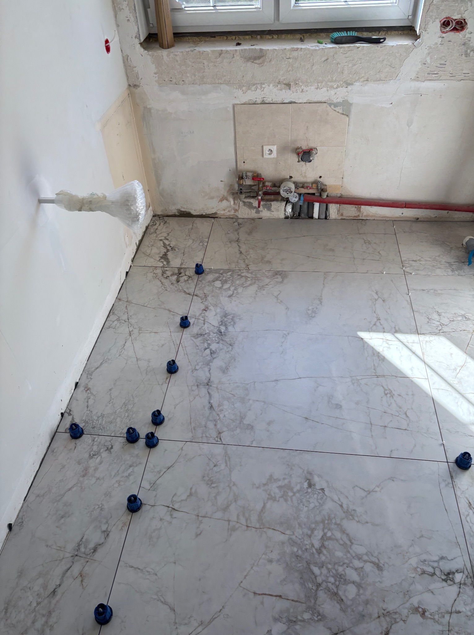 Układanie marmurowych płytek podłogowych z użyciem niebieskich klinów poziomujących, widoczne instalacje w tle, remont kuchni w trakcie.