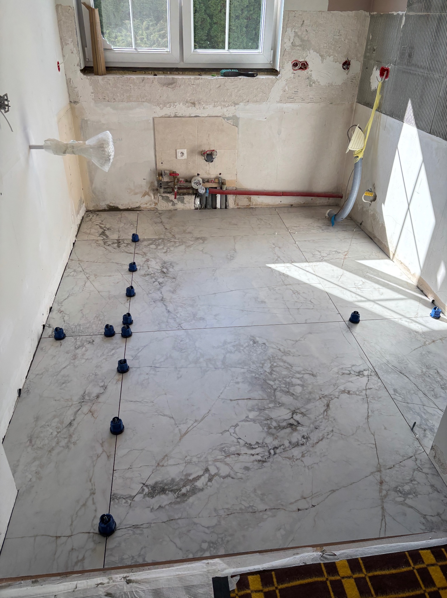 Układanie marmurowych płytek podłogowych w kuchni z użyciem niebieskich klipsów poziomujących. Widoczne rury i okno w tle. Prace wykończeniowe w toku.