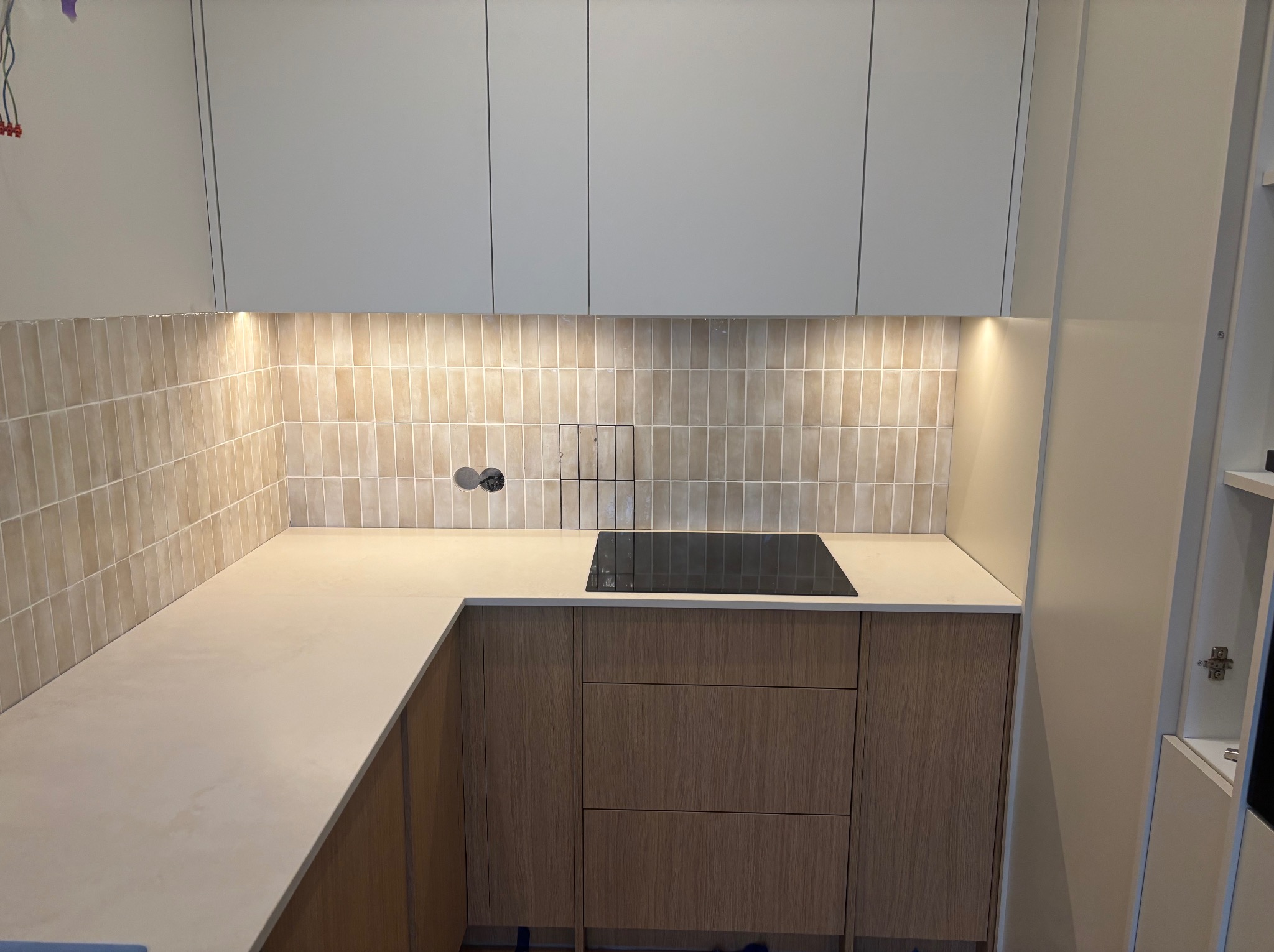 Beżowa kuchnia z drewnianymi szafkami i blatem z konglomeratu. Podświetlane płytki ścienne w odcieniach beżu. Minimalistyczny design z płytą indukcyjną.