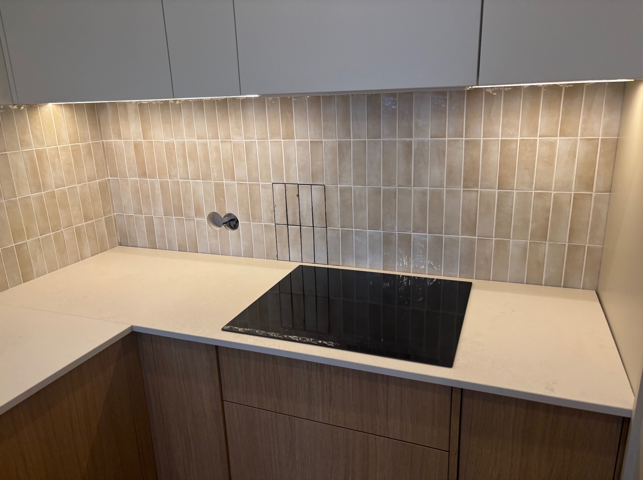 Jasny blat kamienny z płytą indukcyjną w nowoczesnej kuchni, beżowe, pionowe płytki na ścianie, oświetlenie LED podszafkowe. Minimalistyczny design.