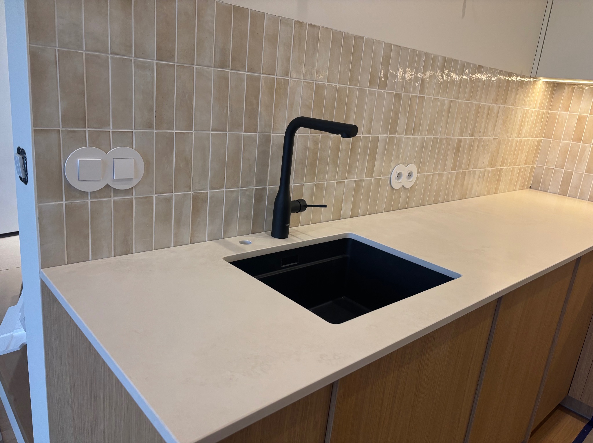 Jasny blat kuchenny z czarnym zlewem i baterią na tle beżowych, pionowo ułożonych płytek. Widoczne gniazdka i włączniki.
