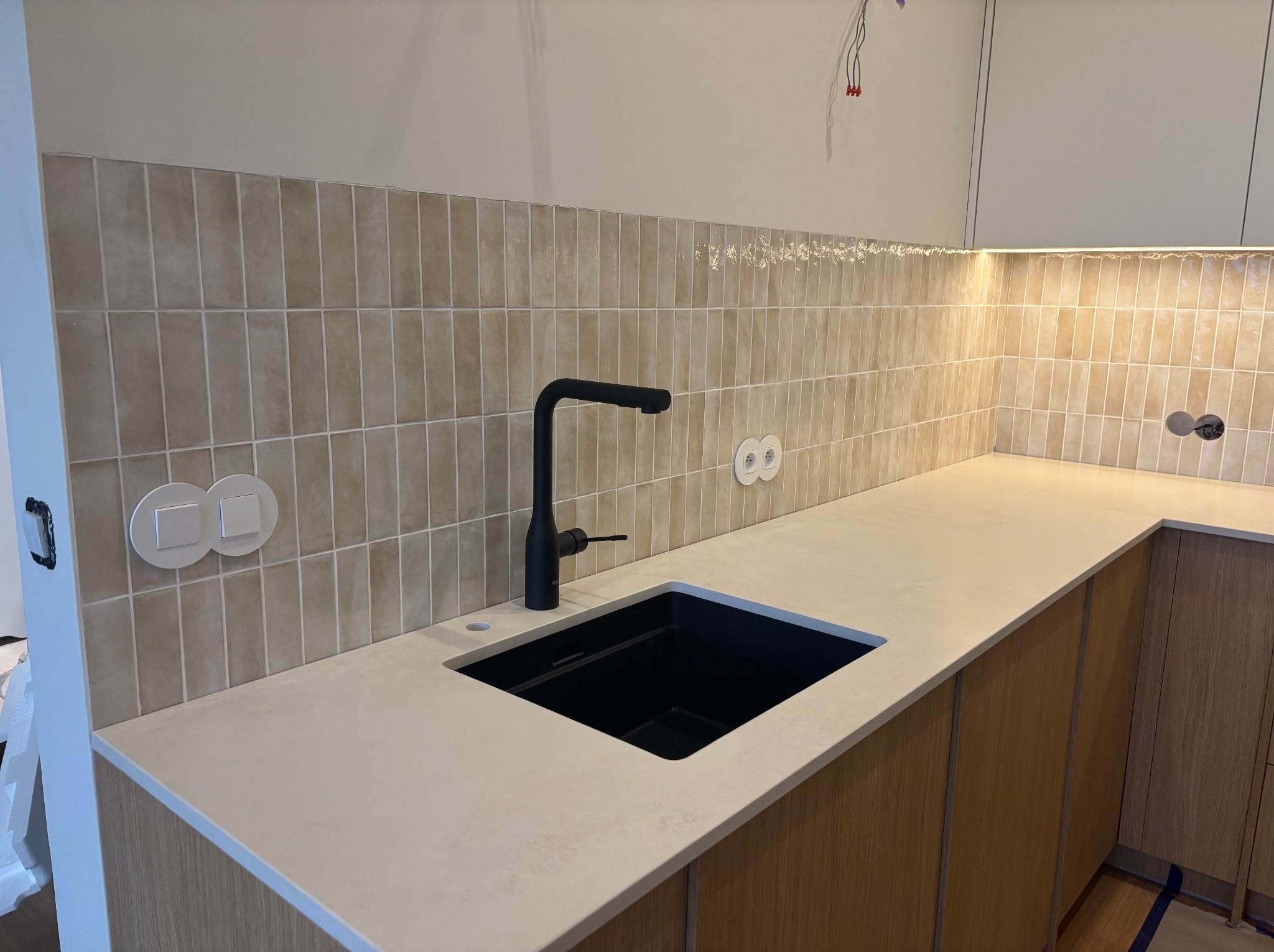 Jasny blat kamienny z czarnym zlewem i baterią w kuchni w Strzelcach Górnych. Beżowe płytki na ścianie i drewniane fronty szafek.