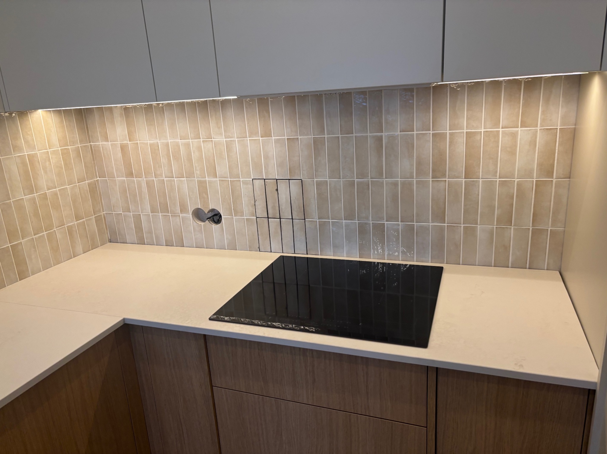 Jasny blat kamienny z płytą indukcyjną w nowoczesnej kuchni, oświetlony ledami, z beżowymi płytkami na ścianie i drewnianymi szafkami.