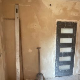 PatPro Bau - Pokój przed remontem: ściany z resztkami farby, zabezpieczone drzwi, poziomica i łopata. Widoczne ślady po poprzednich pracach i przygotowania do szpachlowania.