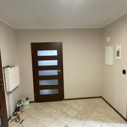 PatPro Bau - Jasne wnętrze z trzema parami drzwi w różnych odcieniach brązu, grzejnikiem i narzędziami malarskimi. Na podłodze płytki w odcieniach beżu.