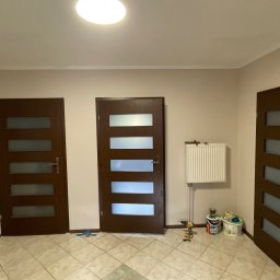 PatPro Bau - Wnętrze z trzema ciemnymi drzwiami z przeszkleniami, grzejnikiem i narzędziami malarskimi. Widok na korytarz po remoncie z nowymi drzwiami.