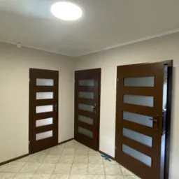 Wnętrze po remoncie: trzy brązowe drzwi z przeszkleniami, kremowe ściany i terakota. Jeden drzwi otwarte, widoczny grzejnik i narzędzia. Oświetlenie sufitowe.