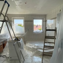 PatPro Bau - Remontowana łazienka z białymi płytkami, zabezpieczona folią malarską. Widoczne dwie drabiny i okna z widokiem na zewnątrz. Przygotowanie do malowania sufitu.