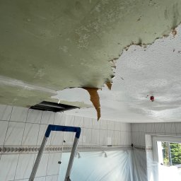 PatPro Bau - Uszkodzony sufit w łazience z widoczną drabiną i zabezpieczoną folią wanną. Fragmenty odpadającego tynku i farby, odsłonięta konstrukcja sufitu.