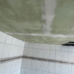 PatPro Bau - Niedokończony remont łazienki: sufit z plamami i częściowo położone płytki. Widoczny fragment drabiny. Ujęcie z dołu, ukazujące stan przed ukończeniem prac.