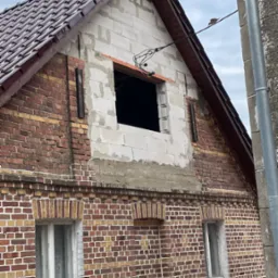 Budynek z cegły z oknem w trakcie montażu, widoczne białe bloczki i elementy konstrukcyjne dachu. Część budynku jest w trakcie prac elewacyjnych.