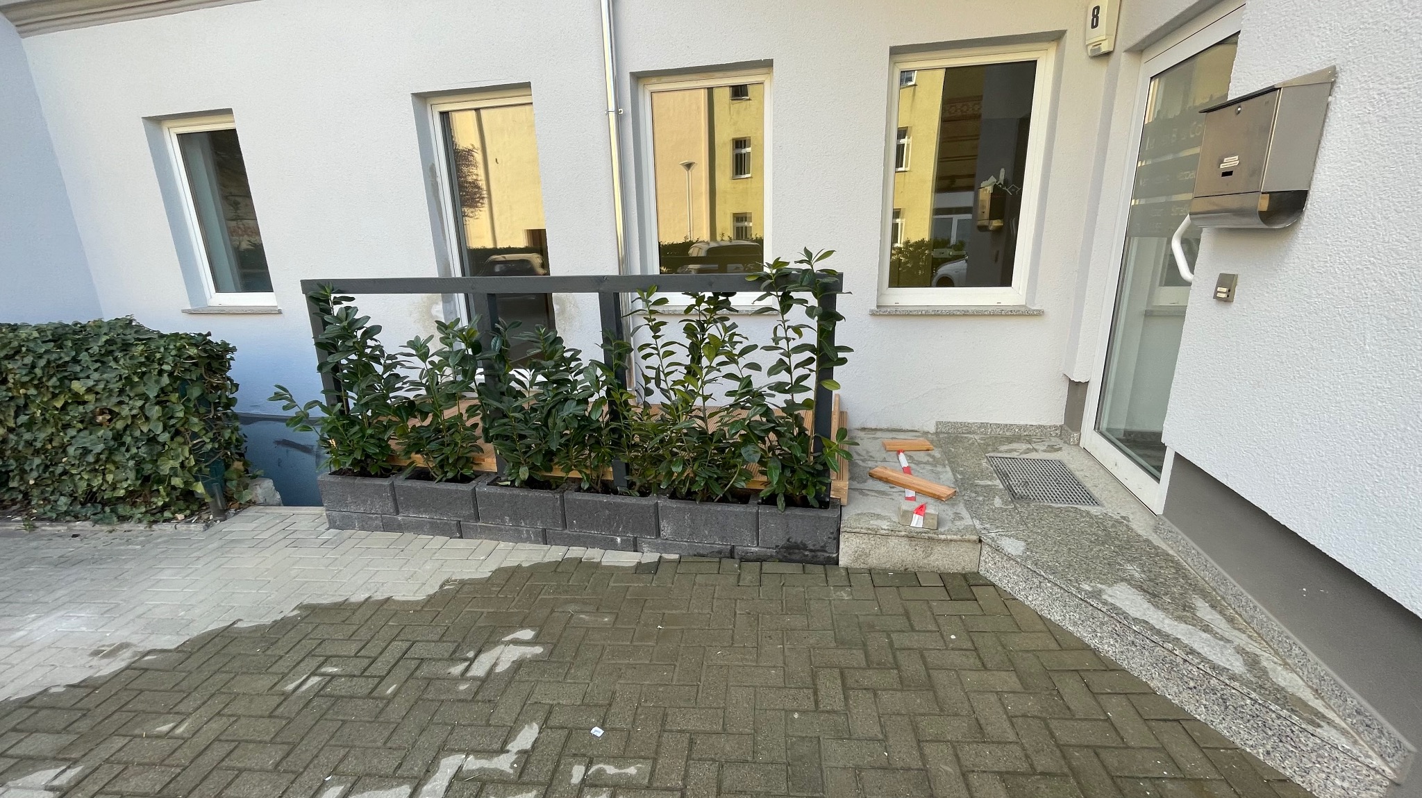 Zewnętrzna zabudowa balkonu z roślinami w donicach z szarej kostki. Na pierwszym planie mokra, brukowa nawierzchnia i fragment zielonego żywopłotu.