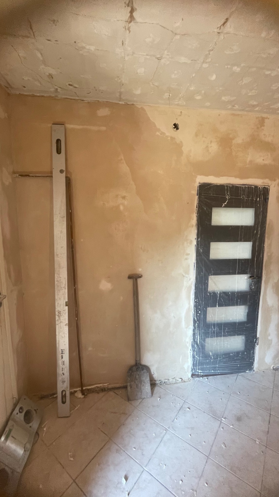 Pokój przed remontem: ściany z resztkami farby, zabezpieczone drzwi, poziomica i łopata. Widoczne ślady po poprzednich pracach i przygotowania do szpachlowania.