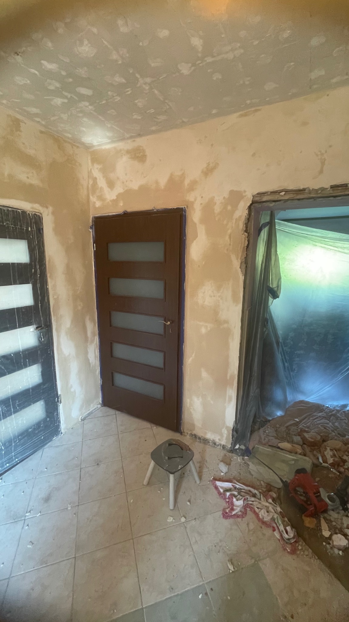 Remont pokoju: ściany po szpachlowaniu, częściowo pomalowane, nowe drzwi z szybami, podłoga z płytek, widoczne narzędzia i gruz. Prace wykończeniowe w trakcie.