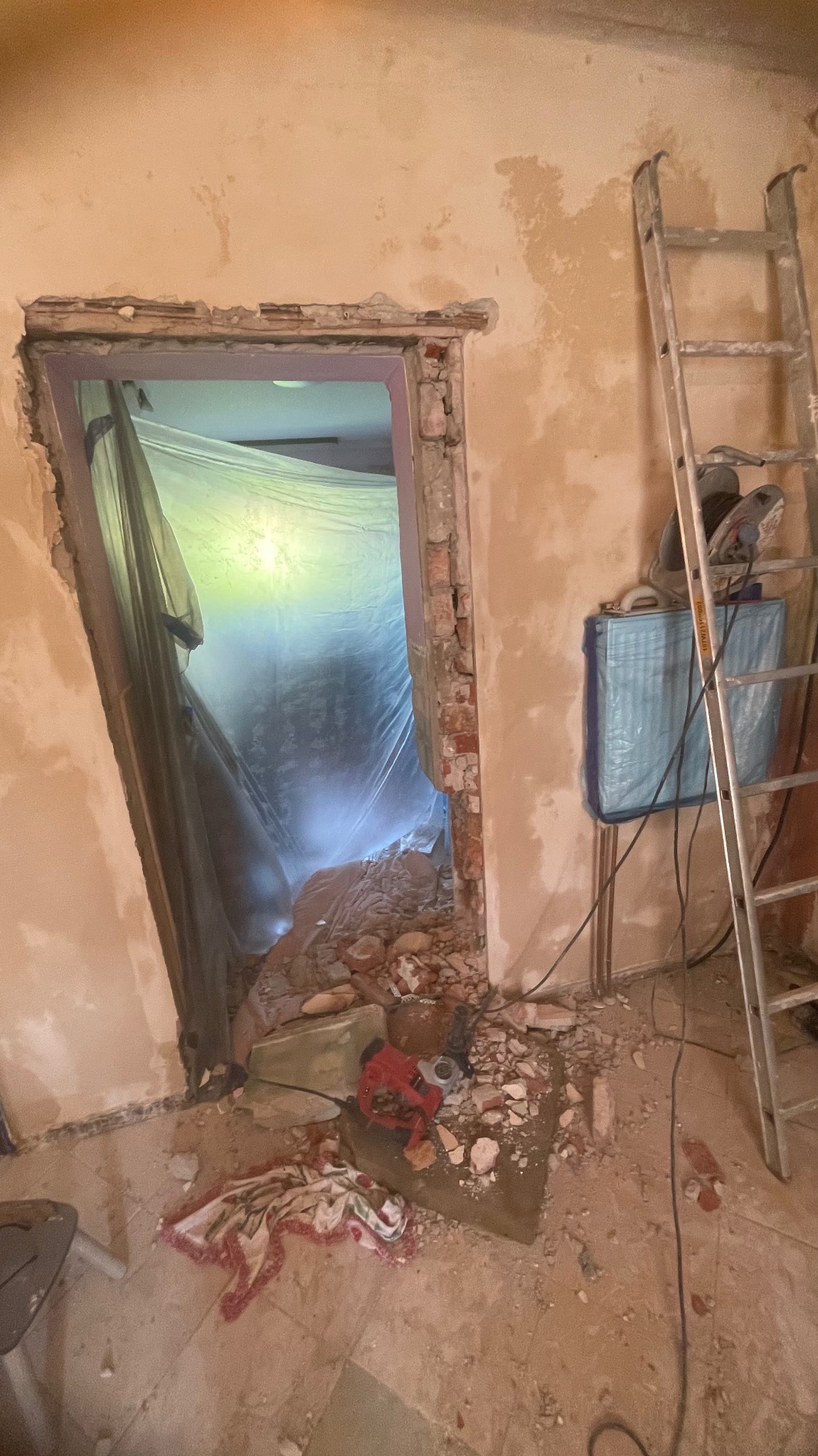 Demontaż ościeżnicy podczas remontu. Widoczne gruz, narzędzia i drabina. Otwór zabezpieczony folią. Ściana z resztkami tapety. Ujęcie z bliska.