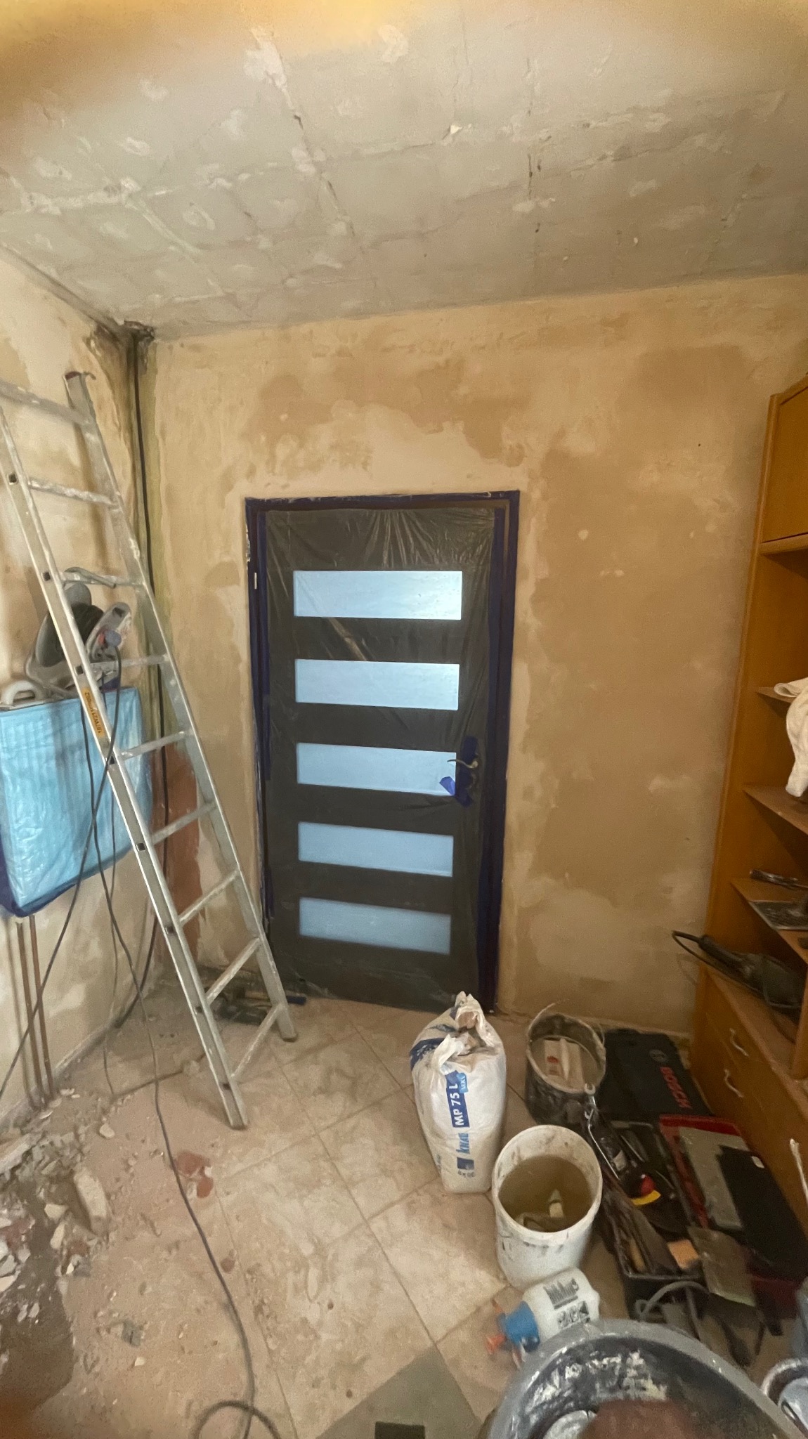 Pokój w trakcie remontu: zabezpieczone folią drzwi, drabina, worki i narzędzia budowlane. Ściany przygotowane do malowania, widoczne ślady szpachlowania.