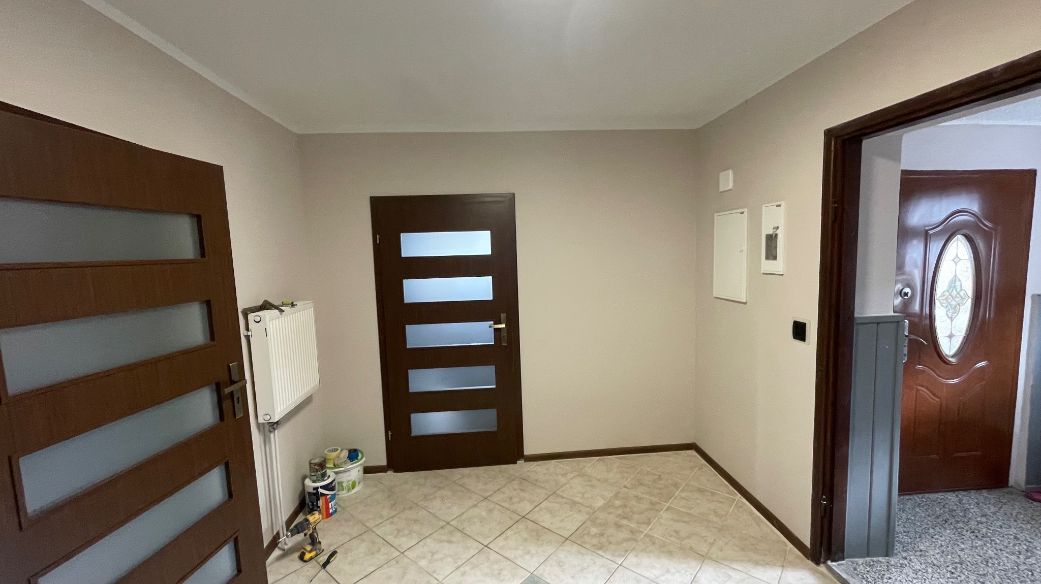 Jasne wnętrze z trzema parami drzwi w różnych odcieniach brązu, grzejnikiem i narzędziami malarskimi. Na podłodze płytki w odcieniach beżu.