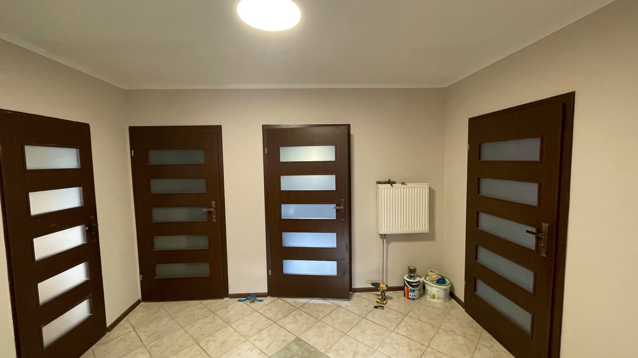 Wnętrze z trzema ciemnymi drzwiami z przeszkleniami, grzejnikiem i narzędziami malarskimi. Widok na korytarz po remoncie z nowymi drzwiami.