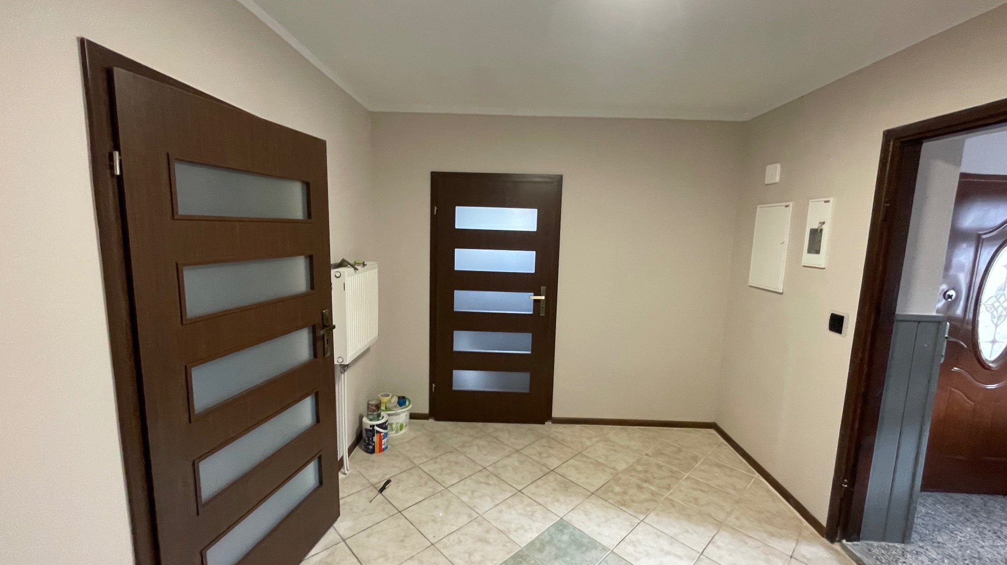 Korytarz z brązowymi drzwiami z przeszkleniami i białym grzejnikiem. Na podłodze płytki w odcieniach beżu. Widoczne puszki po farbach.