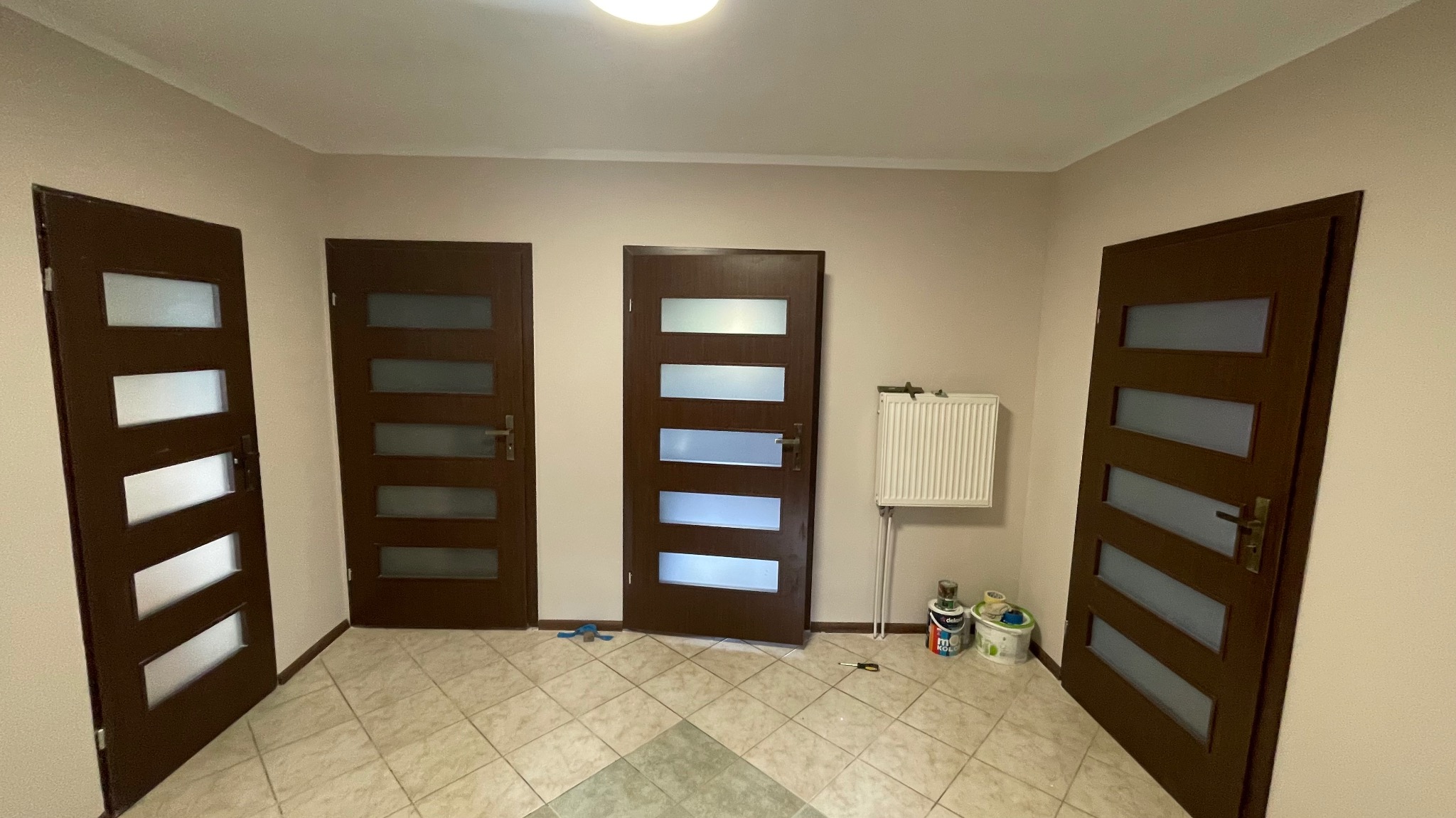 Wnętrze z trzema zamontowanymi, brązowymi drzwiami z szybami, grzejnikiem i akcesoriami malarskimi na podłodze z płytek. Widok na korytarz po remoncie.