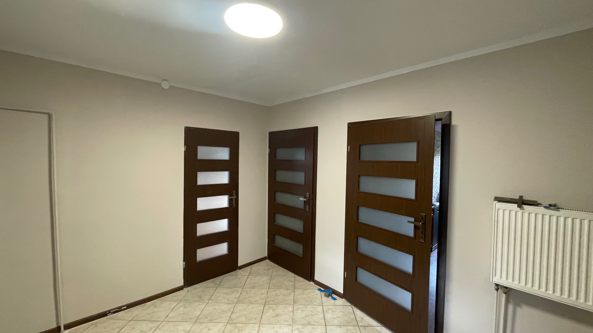 Wnętrze po remoncie: trzy brązowe drzwi z przeszkleniami, kremowe ściany i terakota. Jeden drzwi otwarte, widoczny grzejnik i narzędzia. Oświetlenie sufitowe.