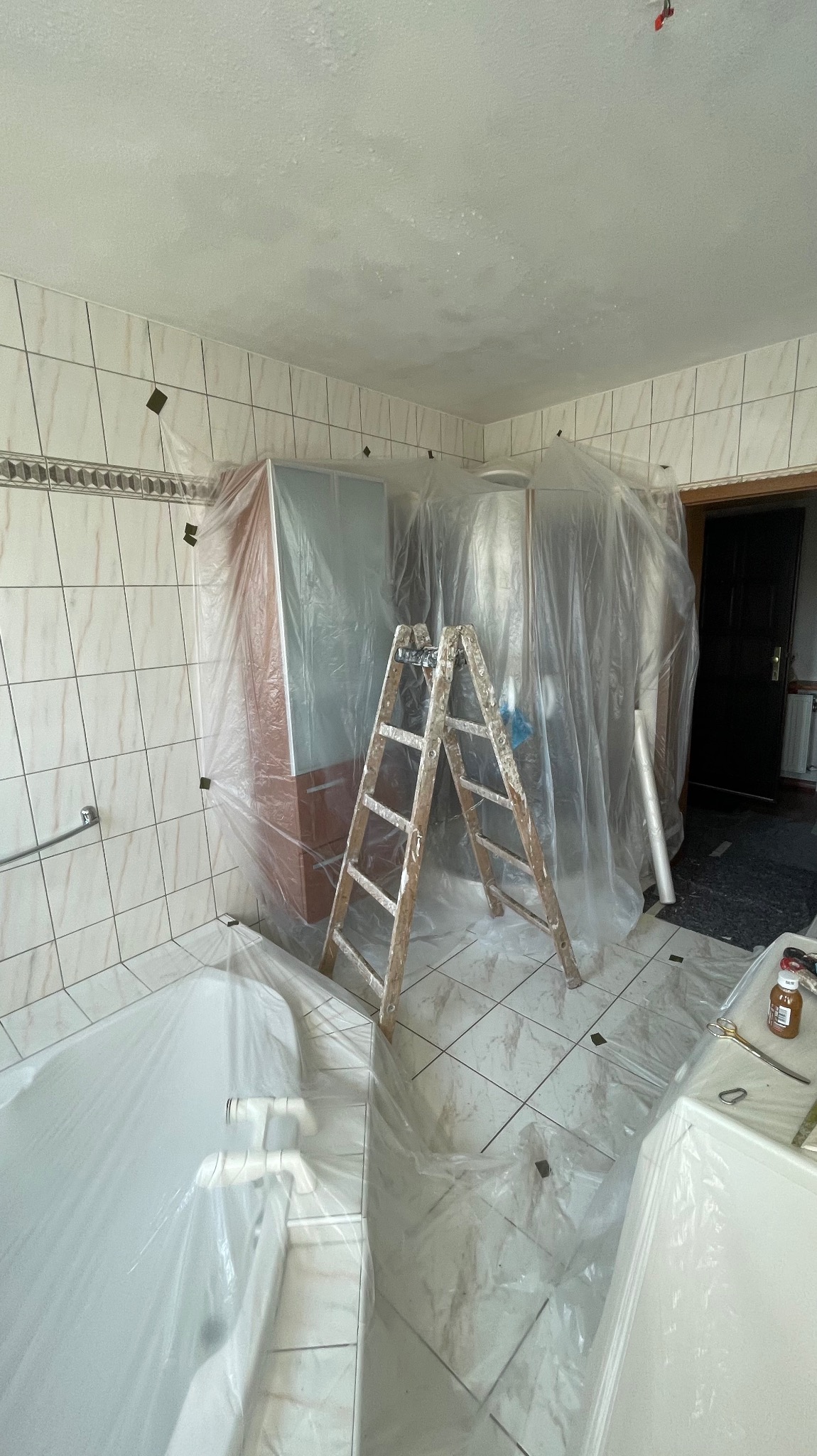 Łazienka w trakcie remontu: zabezpieczone folią meble, wanna i podłoga. Stara, drewniana drabina oparta o ścianę. Przygotowanie do malowania sufitu.