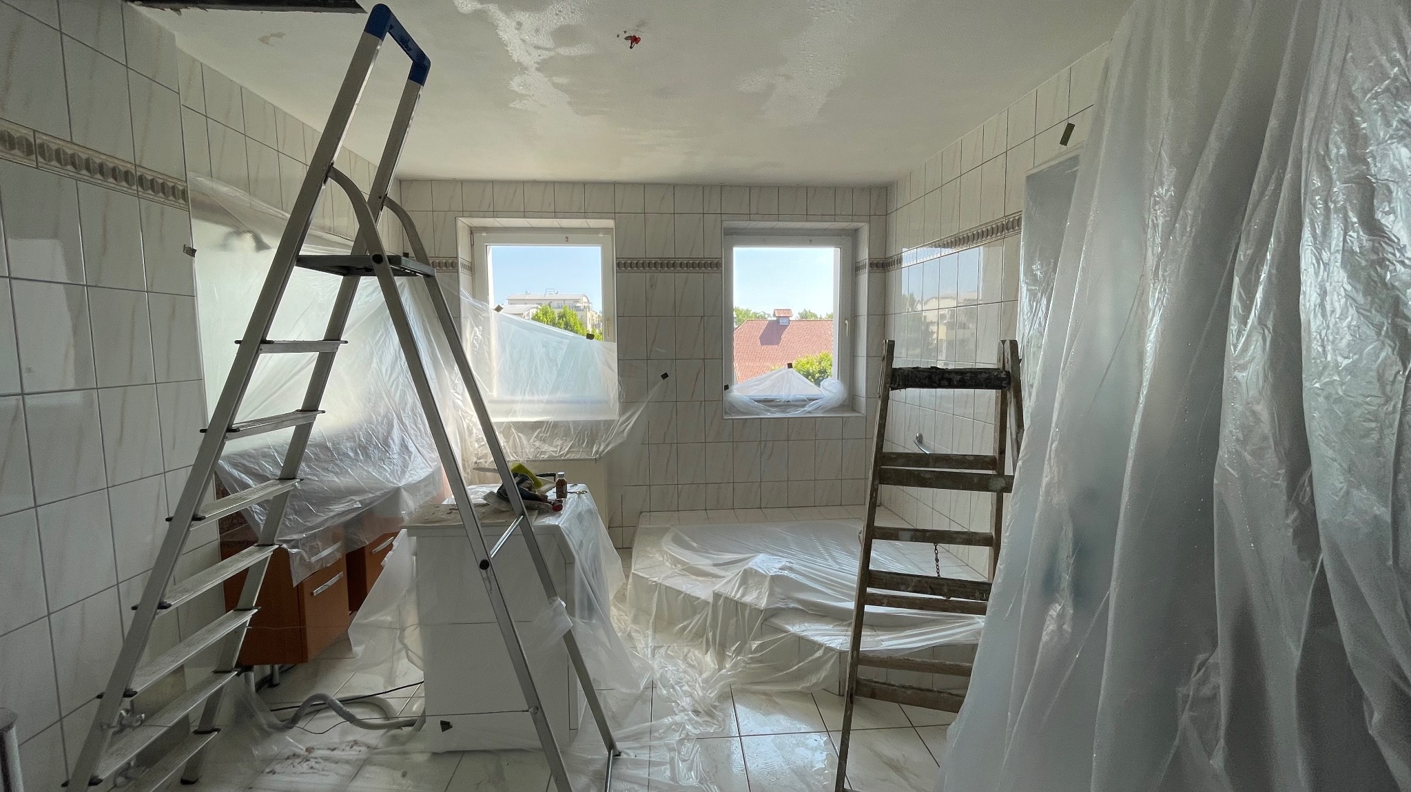 Remontowana łazienka z białymi płytkami, zabezpieczona folią malarską. Widoczne dwie drabiny i okna z widokiem na zewnątrz. Przygotowanie do malowania sufitu.