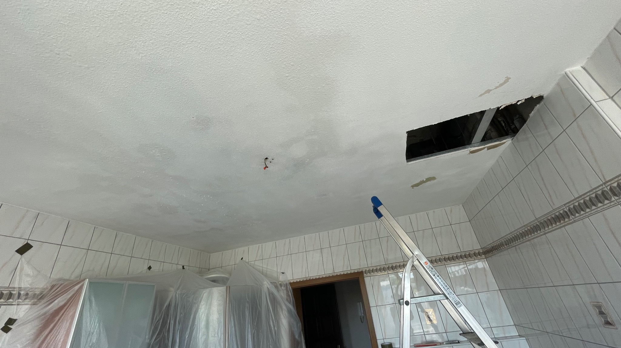 Biały sufit z otworem i śladami szpachlowania w pomieszczeniu wyłożonym białymi płytkami. Aluminiowa drabina oparta o ścianę. Prace przygotowawcze do remontu.
