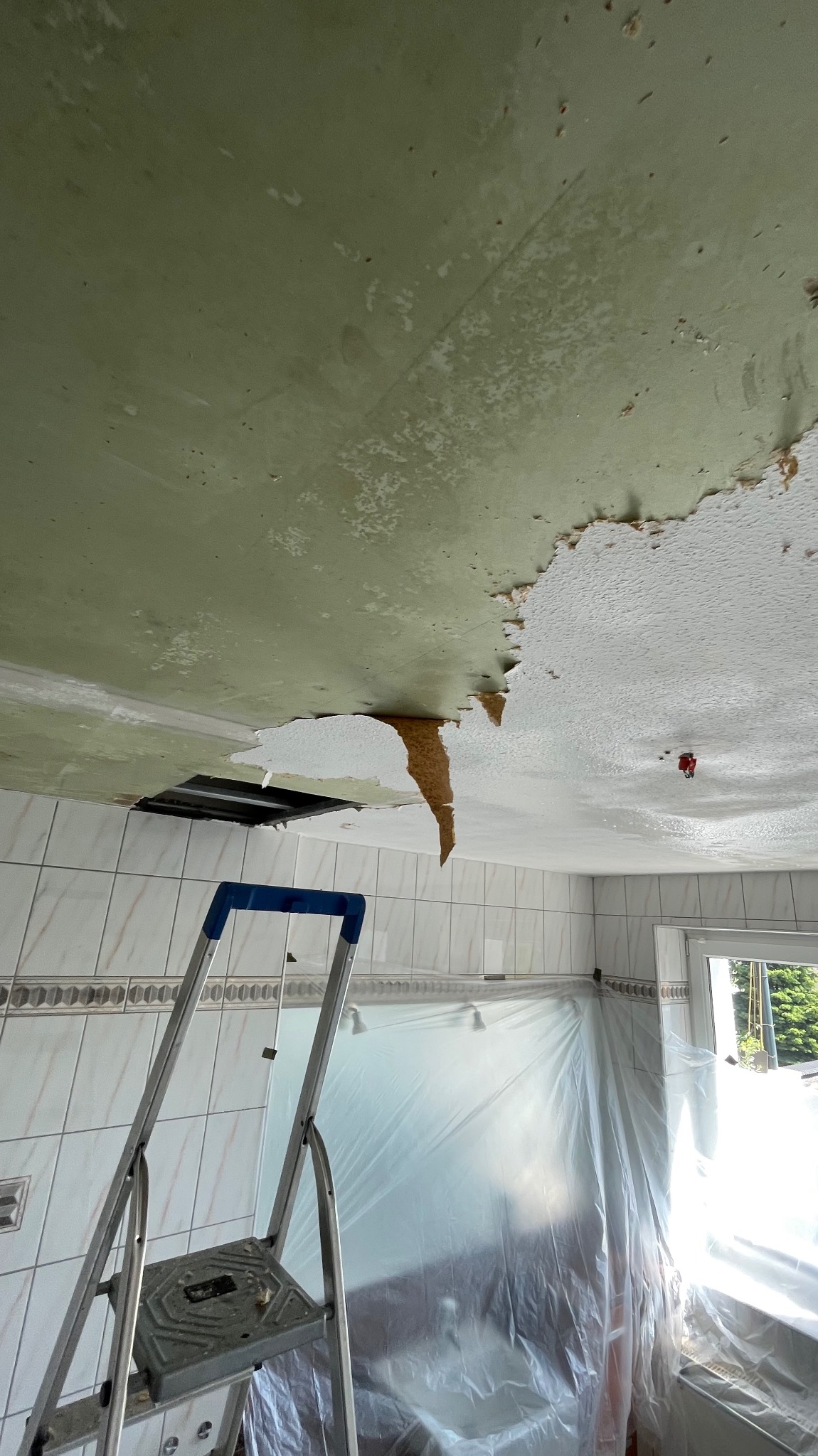 Uszkodzony sufit w łazience z widoczną drabiną i zabezpieczoną folią wanną. Fragmenty odpadającego tynku i farby, odsłonięta konstrukcja sufitu.