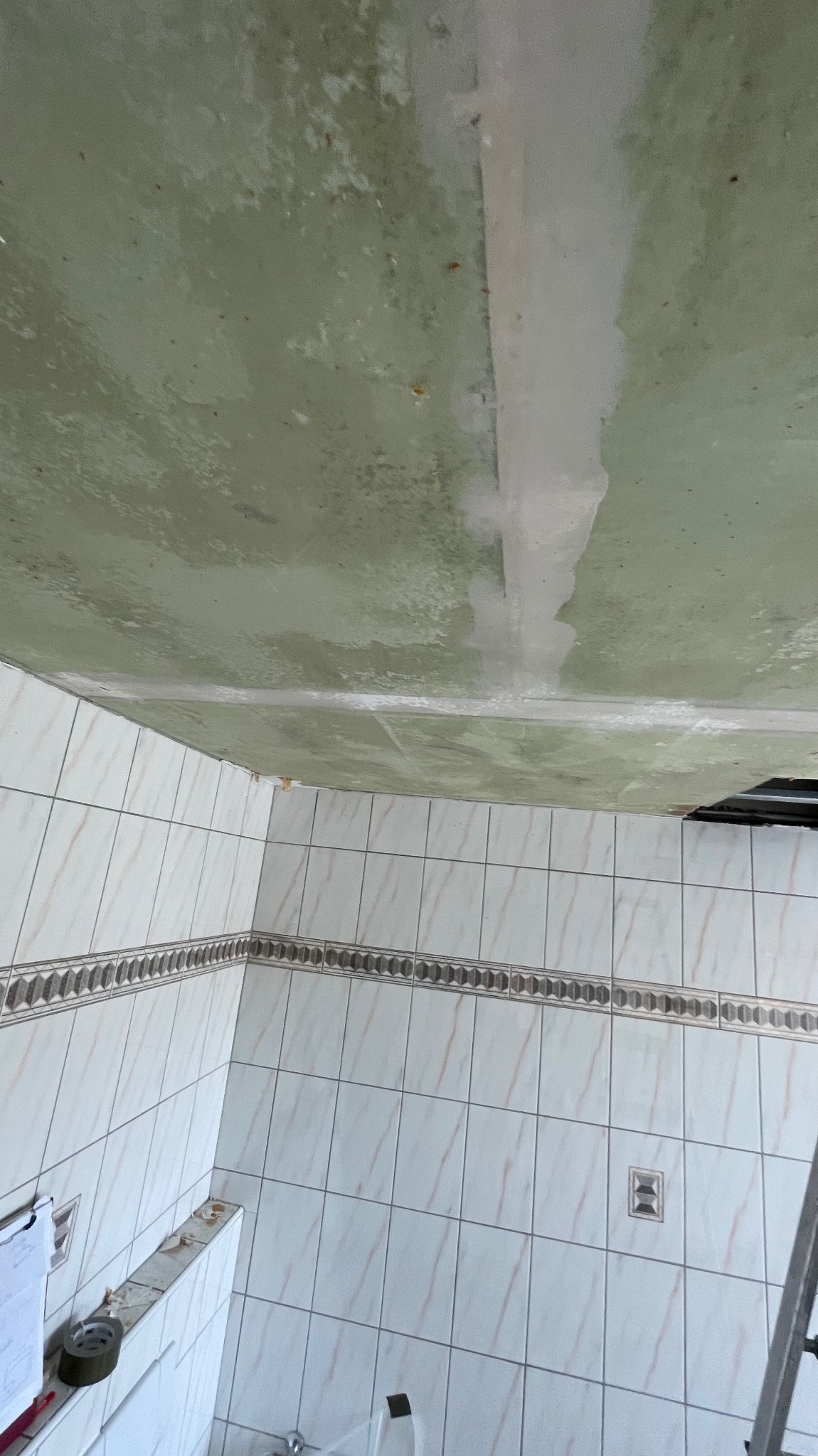 Niedokończony remont łazienki: sufit z plamami i częściowo położone płytki. Widoczny fragment drabiny. Ujęcie z dołu, ukazujące stan przed ukończeniem prac.