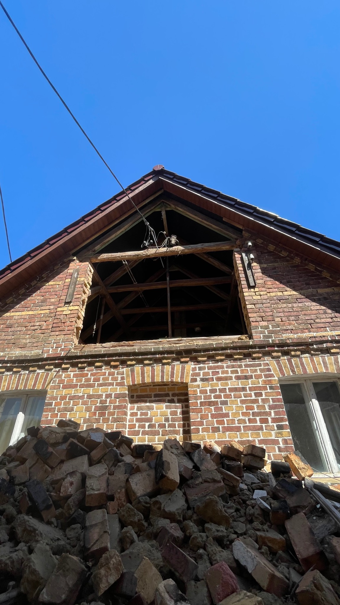 Ceglany budynek w trakcie rozbiórki, z widocznym otworem w dachu i stertą gruzu u podstawy, sfotografowany pod kątem w słoneczny dzień.