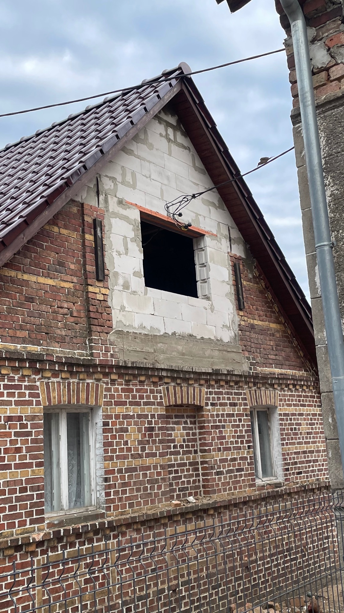 Budynek z cegły z oknem w trakcie montażu, widoczne białe bloczki i elementy konstrukcyjne dachu. Część budynku jest w trakcie prac elewacyjnych.