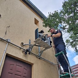 KLIMAKOS - Mężczyzna na drabinie serwisuje klimatyzator na elewacji budynku, widoczne rury i elementy instalacji, drzewo w tle.