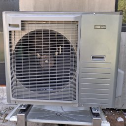 KLIMAKOS - Szara zewnętrzna jednostka pompy ciepła Nibe AMS20 na wspornikach, ustawiona przy szarej ścianie budynku. Widoczna kratka wentylatora i logo producenta.