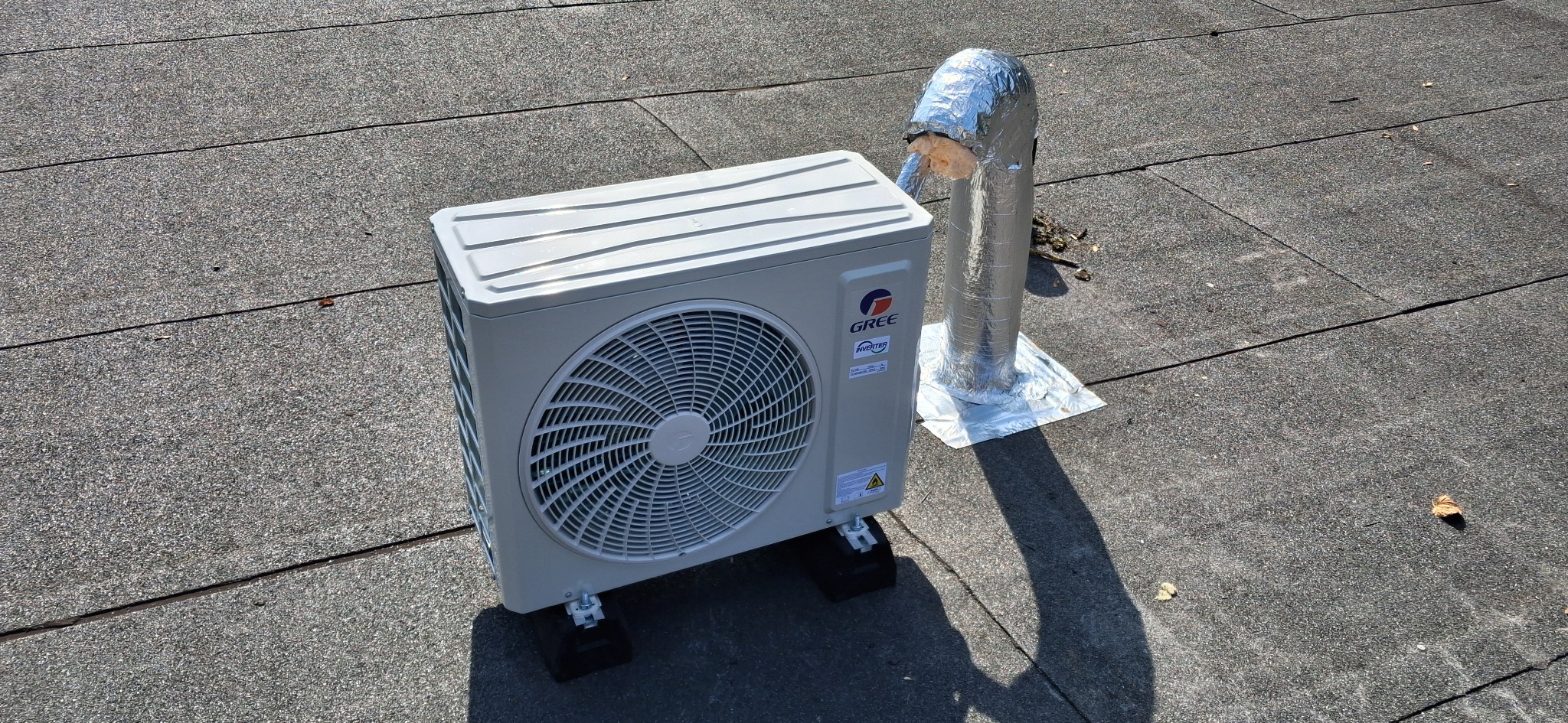 Biała jednostka zewnętrzna klimatyzacji Gree na czarnym dachu z papy, obok srebrna rura wentylacyjna z izolacją. Widoczne czarne podkładki pod urządzeniem.