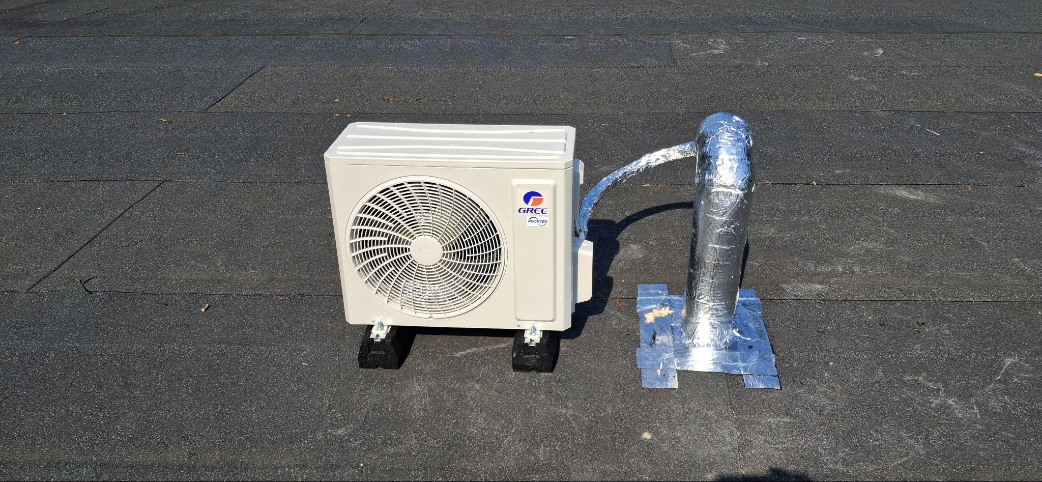 Klimatyzator marki Gree na dachu pokrytym papą, z widocznym odprowadzeniem skroplin owiniętym folią aluminiową. Widok z boku, w słoneczny dzień.