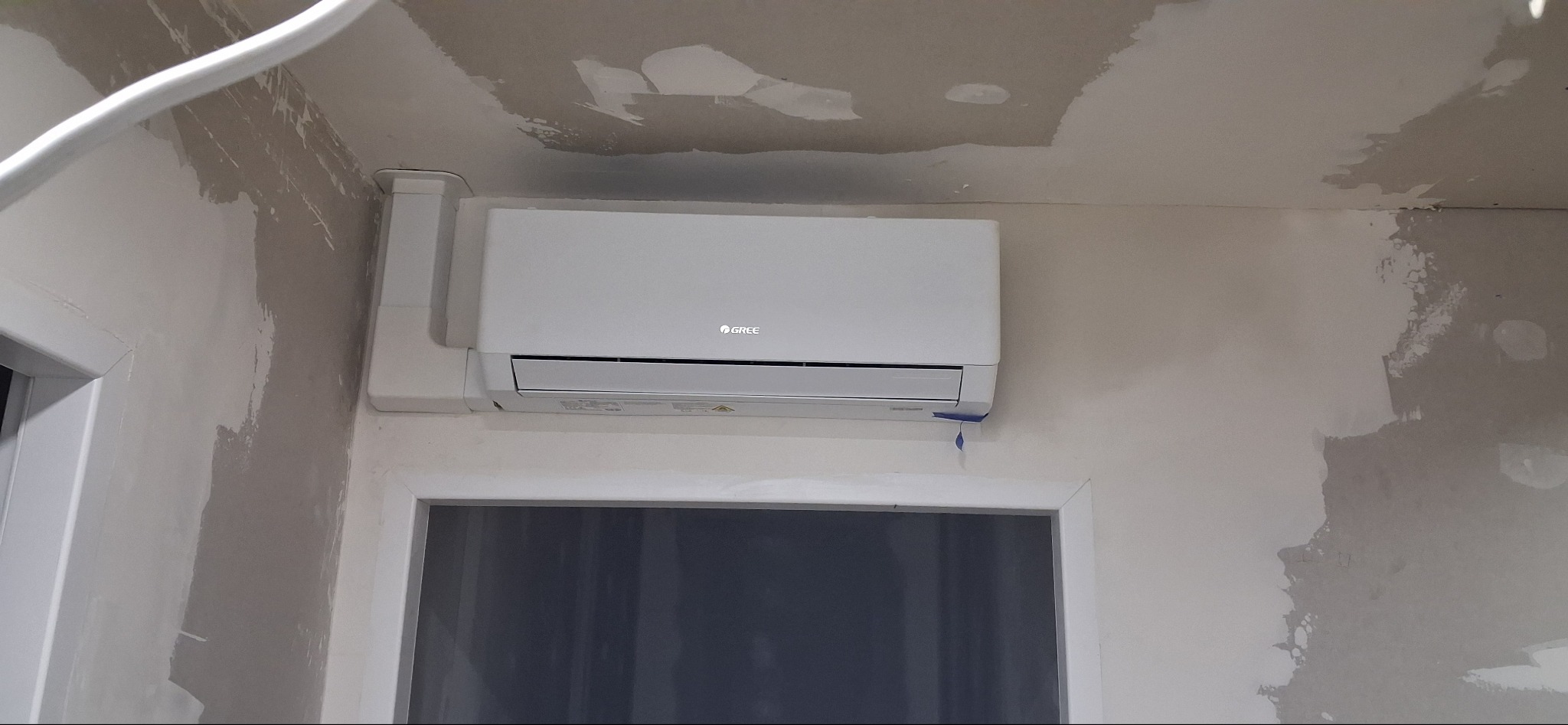 Biały klimatyzator Gree zamontowany na ścianie w trakcie wykańczania, widoczne ślady po gładzi. Instalacja prowadzona w listwach.