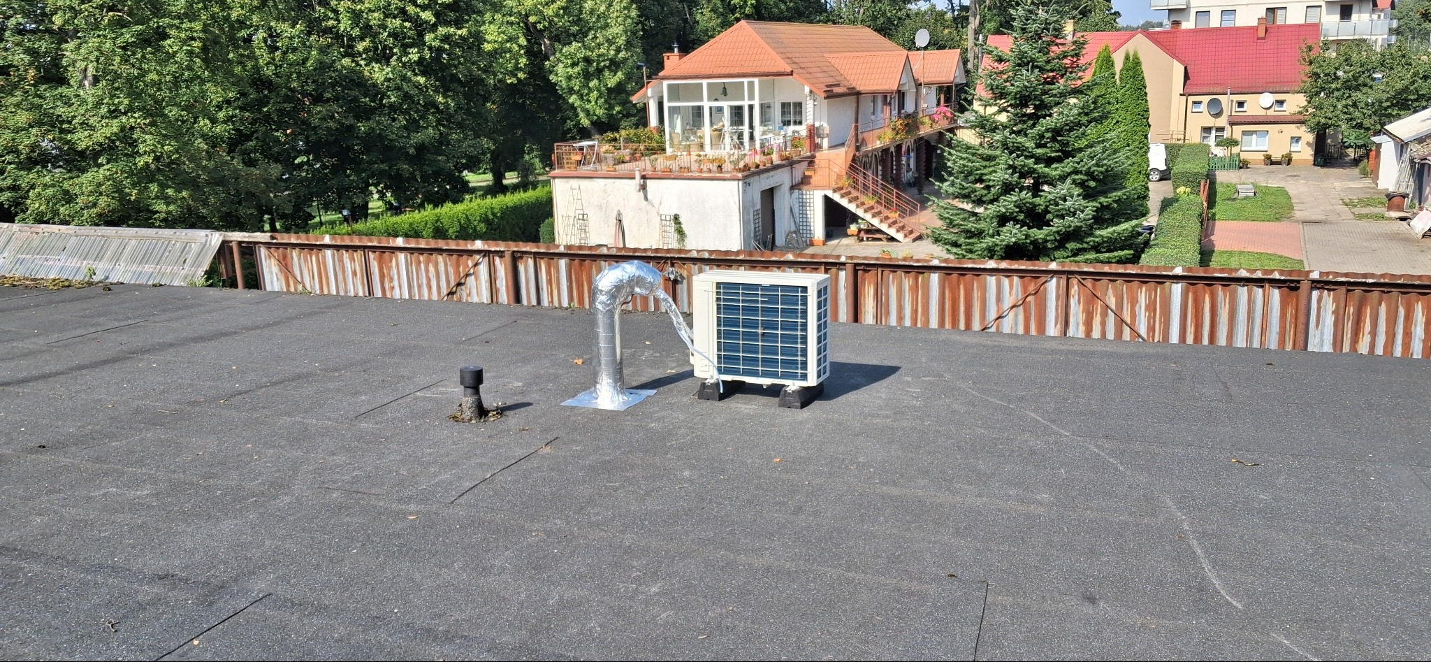 Kompaktowa jednostka zewnętrzna klimatyzacji na płaskim dachu budynku, z widocznym przewodem i kominem wentylacyjnym. W tle zabudowa jednorodzinna i zieleń.